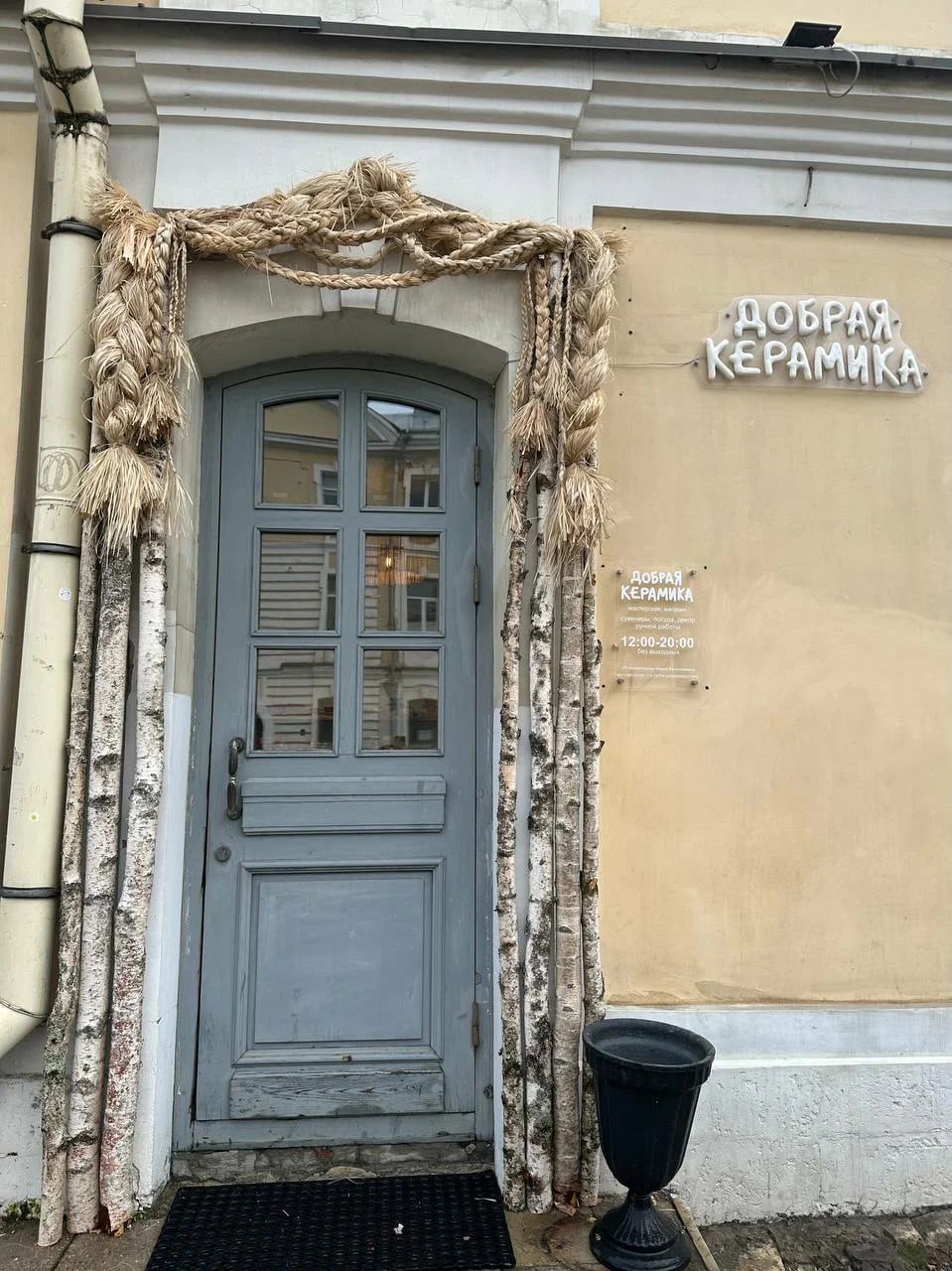 Вход в мастерскую «Добрая керамика»: арочная дверь, убранная плетёными жгутами и берёзовыми стволами, рядом вывеска и табличка с часами работы 12:00–20:00.